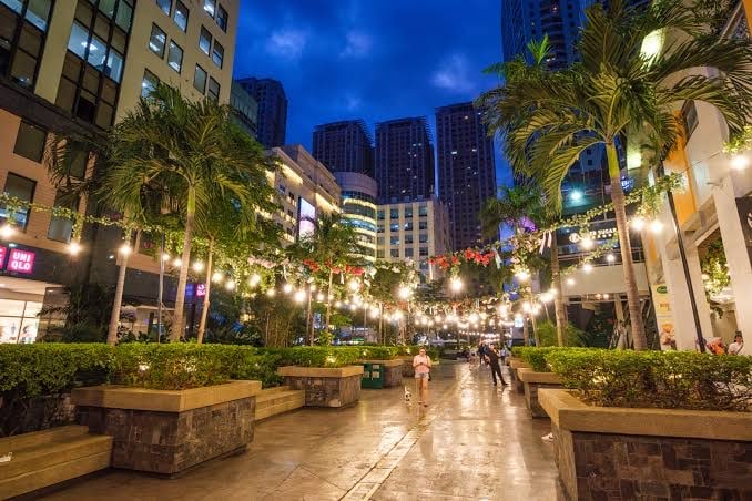 An outdoor pathway is illuminated by string lights, creating a warm ambiance amidst late evening shades of blue. Lush greenery and palm trees line the walkway, leading to modern buildings and a lively plaza. Individuals stroll leisurely, enjoying the surrounding atmosphere.