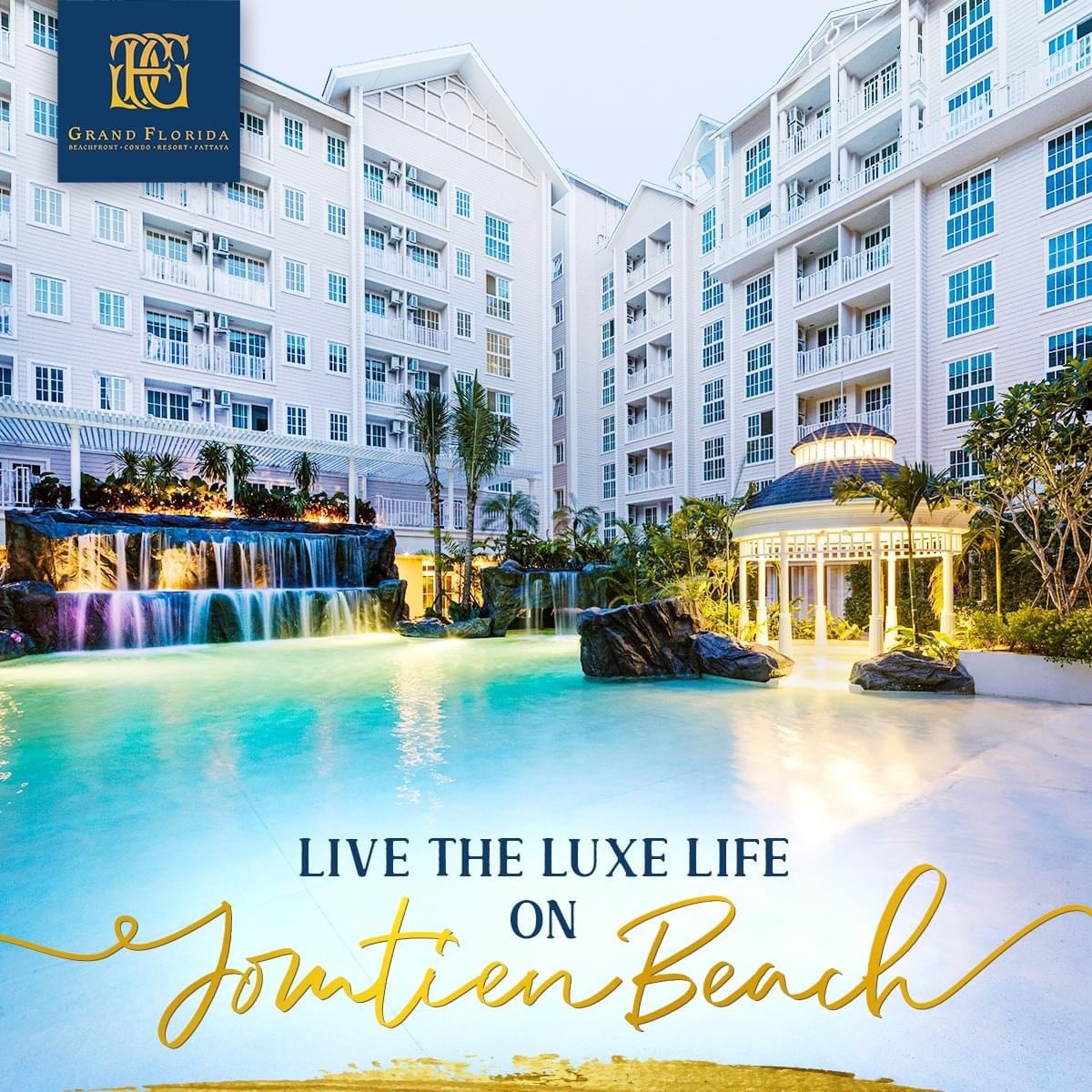 A large, inviting pool area is surrounded by stylized tropical landscaping and elegant architecture. Water features create a serene ambiance, while lounge chairs are strategically placed for relaxation. The building's exterior reflects a blend of modern and classic design elements, offering a welcoming atmosphere.