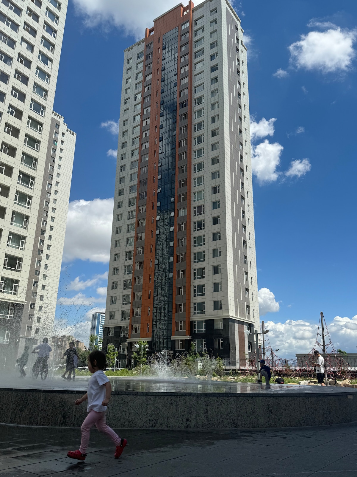 A tall residential building reaches into the sky, featuring a blend of white and orange facade. In the foreground, children are seen playing near a stone fountain with water spraying gently, under a bright blue sky scattered with fluffy white clouds.