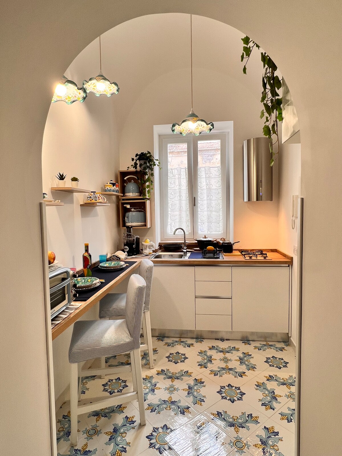 A compact kitchen features modern appliances, including a microwave and gas stove. The space is adorned with decorative tile flooring and hanging light fixtures, creating a bright atmosphere. A small dining area with two stools is positioned near a window that allows natural light to enter.