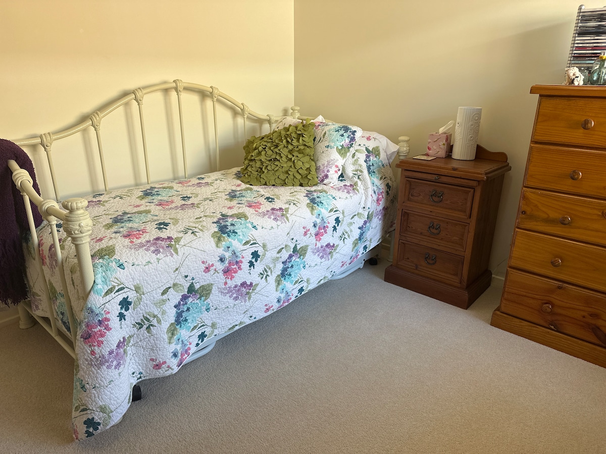 A bright sleeping area features a single bed adorned with a floral quilt. A decorative pillow adds a touch of color, while a wooden bedside table and a tall dresser provide storage options. Soft carpet lines the floor, contributing to the cozy ambiance.