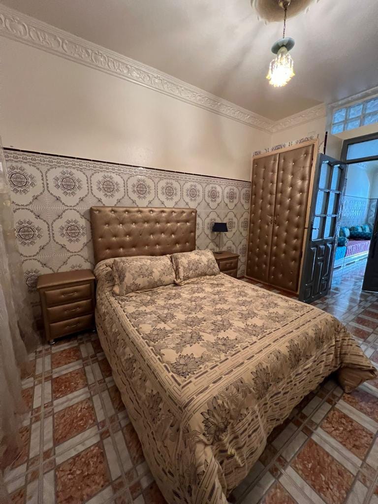 A spacious sleeping area is furnished with a large upholstered bed, complemented by two nightstands on either side. The wall features a decorative patterned design. Light enters through an open doorway, revealing a glimpse of the adjoining room's vibrant decor.