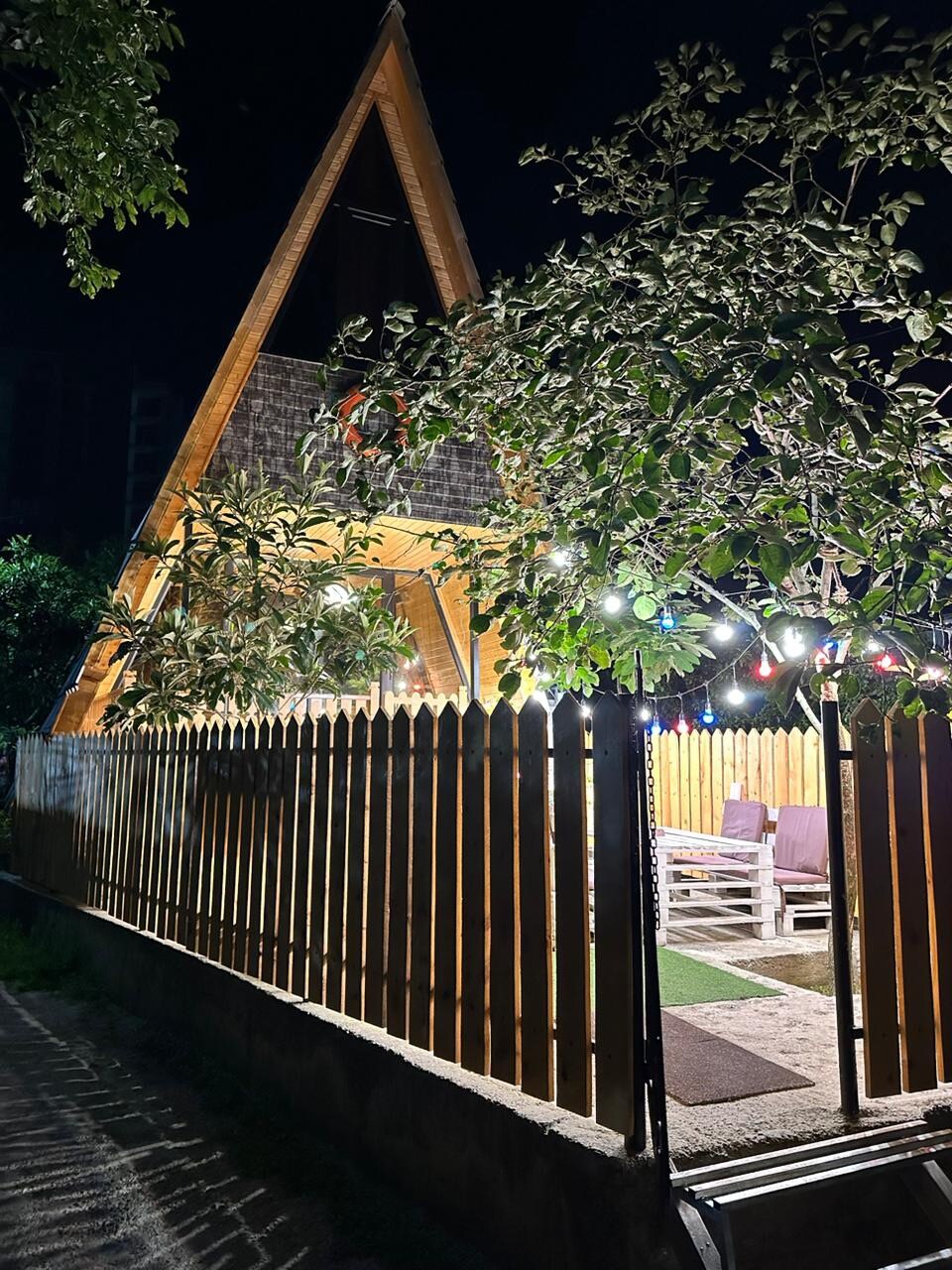 The A-frame structure is illuminated at night by warm outdoor lights, highlighting its unique architectural design. A wooden fence surrounds the property, and lush greenery is visible along the pathway. Cozy seating areas can be seen within the enclosed yard, enhancing the inviting atmosphere.