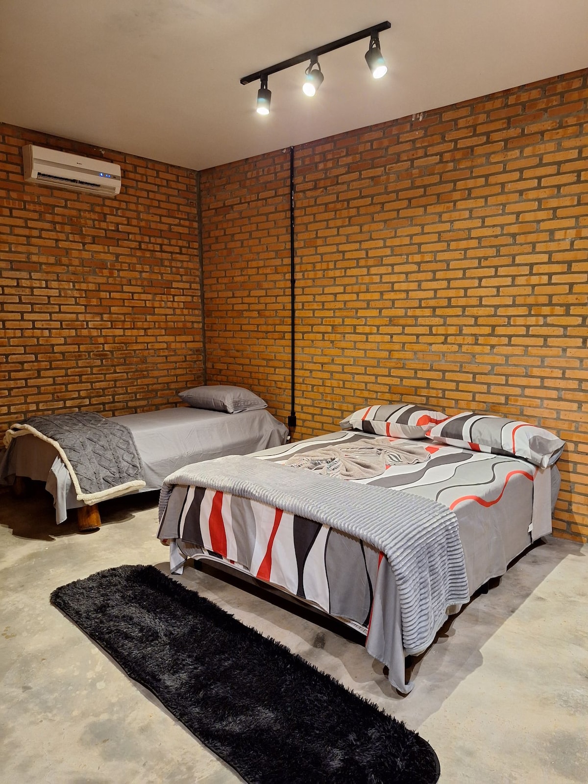 A cozy sleeping area is present, featuring a spacious bed with patterned bedding alongside a smaller bed. Exposed brick walls add texture, while a soft rug lies underfoot. Recessed lighting provides illumination, and a wall-mounted air conditioning unit is positioned for comfort.