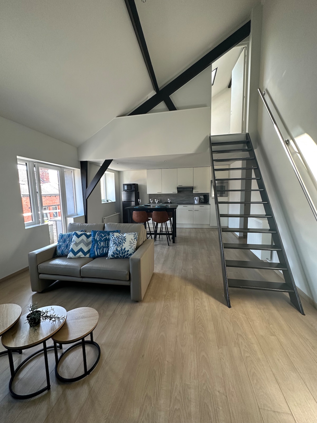 The image presents a modern duplex apartment with a light-filled living area. A cozy sofa adorned with decorative pillows is positioned next to a set of small tables. The open layout reveals a staircase leading to the upper level, with a kitchen area visible in the background.