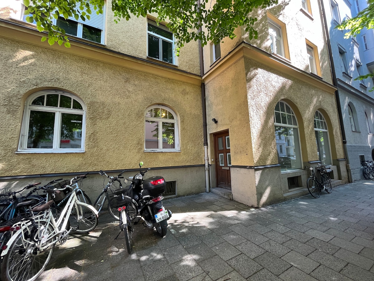 The exterior of a multi-story building is visible, featuring large arched windows and a welcoming entrance. Bicycles are parked along the side, while greenery from overhanging trees provides natural shade. The surrounding area is paved, adding to the urban atmosphere.