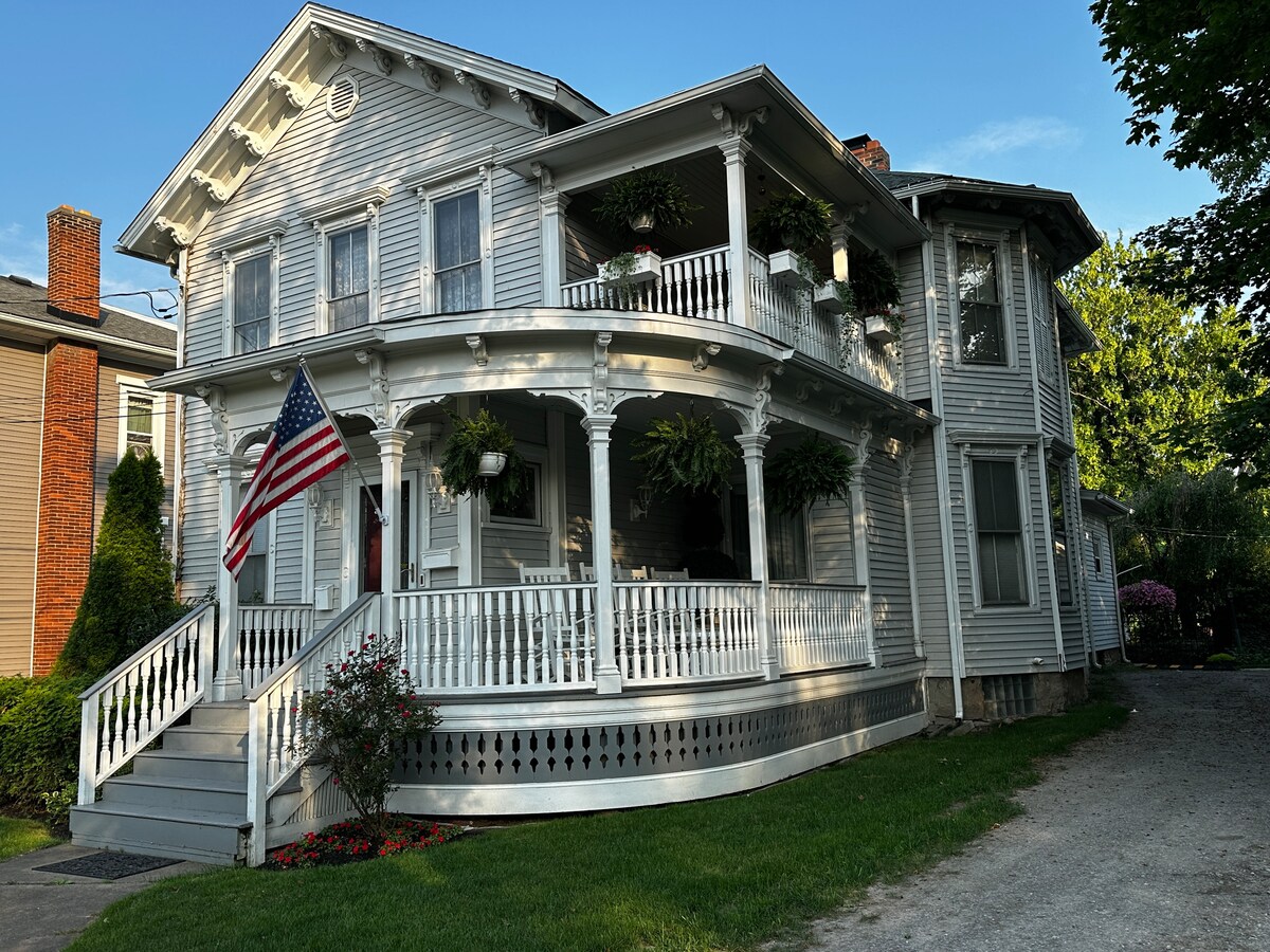 The exterior of a historic, multi-story house features a large and inviting covered porch adorned with hanging plants. An American flag is displayed prominently. Lush green grass surrounds the property, and the architectural details illustrate classic charm and character.