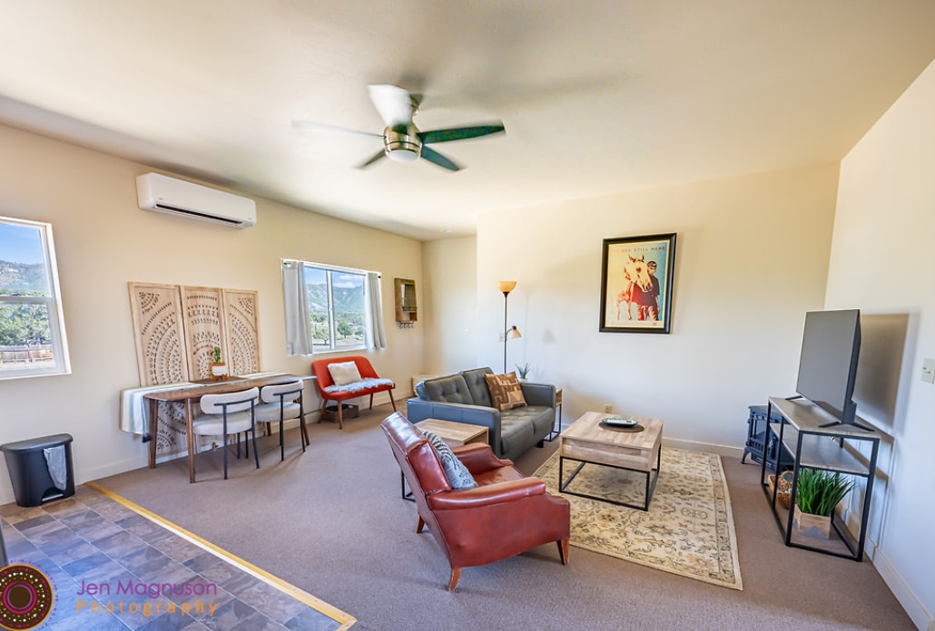 A spacious living area showcases modern furnishings, including a gray sofa and red accent chairs. Natural light fills the room through large windows, revealing scenic views. A wooden coffee table sits in front of a TV on a stand, while a dining table is located nearby.