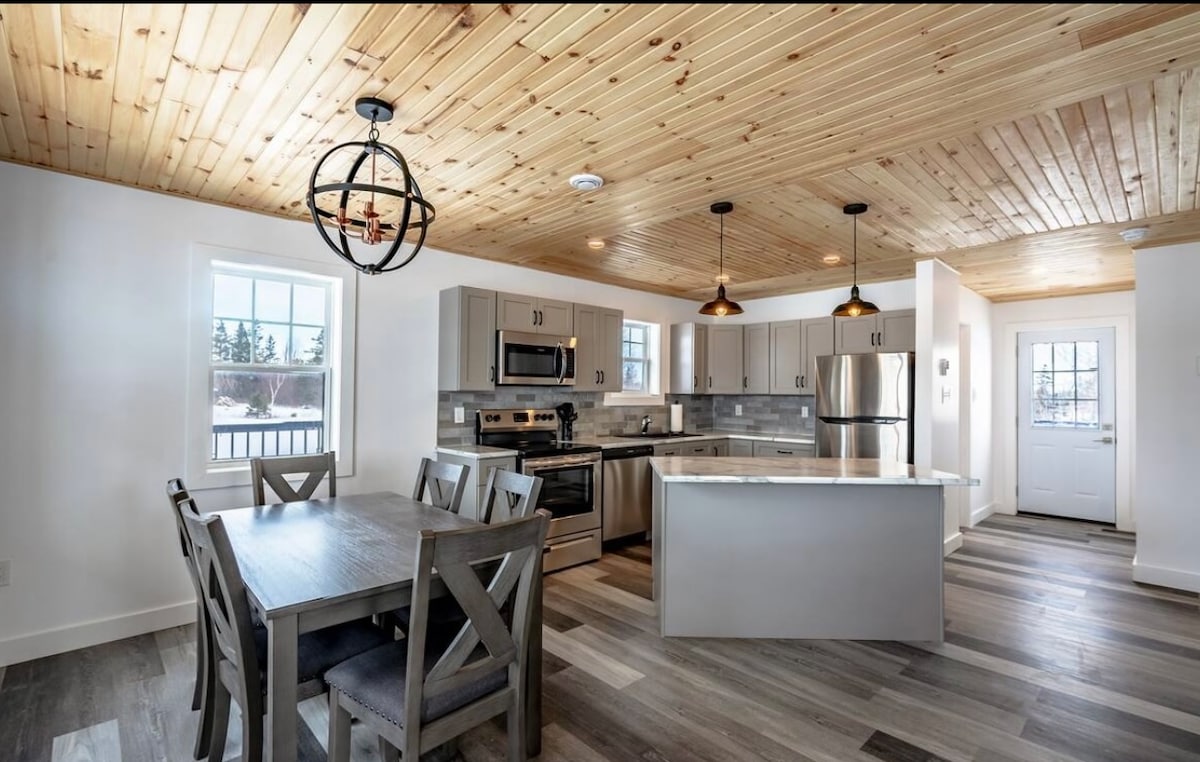 The kitchen is equipped with modern appliances, including a stainless steel refrigerator and stove. A central island offers additional counter space, and a wooden dining table accommodates six. Natural light fills the space through multiple windows, highlighting the wooden ceiling and sleek cabinetry.