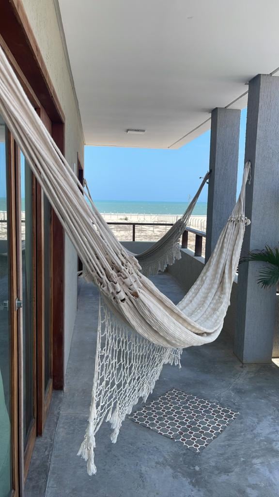 Two hammocks are suspended on a spacious balcony, providing a relaxing area with views of the sea. The flooring is made of concrete, and a patterned mat is placed near the entry. Bright sky and ocean are visible in the background.