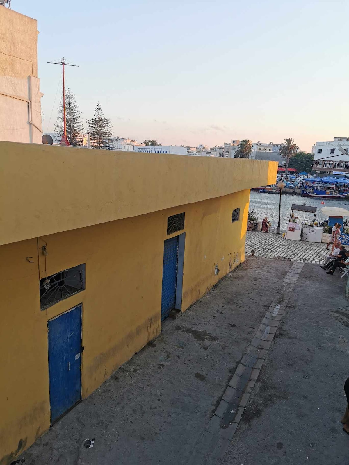 A vibrant yellow building is captured from an elevated angle, featuring blue doors and windows. The surrounding area includes a cobblestone walkway and a view of nearby boats along the waterfront. Soft colors of twilight illuminate the scene.