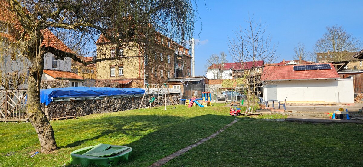 A spacious backyard is presented, featuring a well-maintained lawn with a few trees. Various play equipment and colorful toys are scattered across the area. A covered structure and a building with solar panels are seen in the background, contributing to the overall outdoor environment.