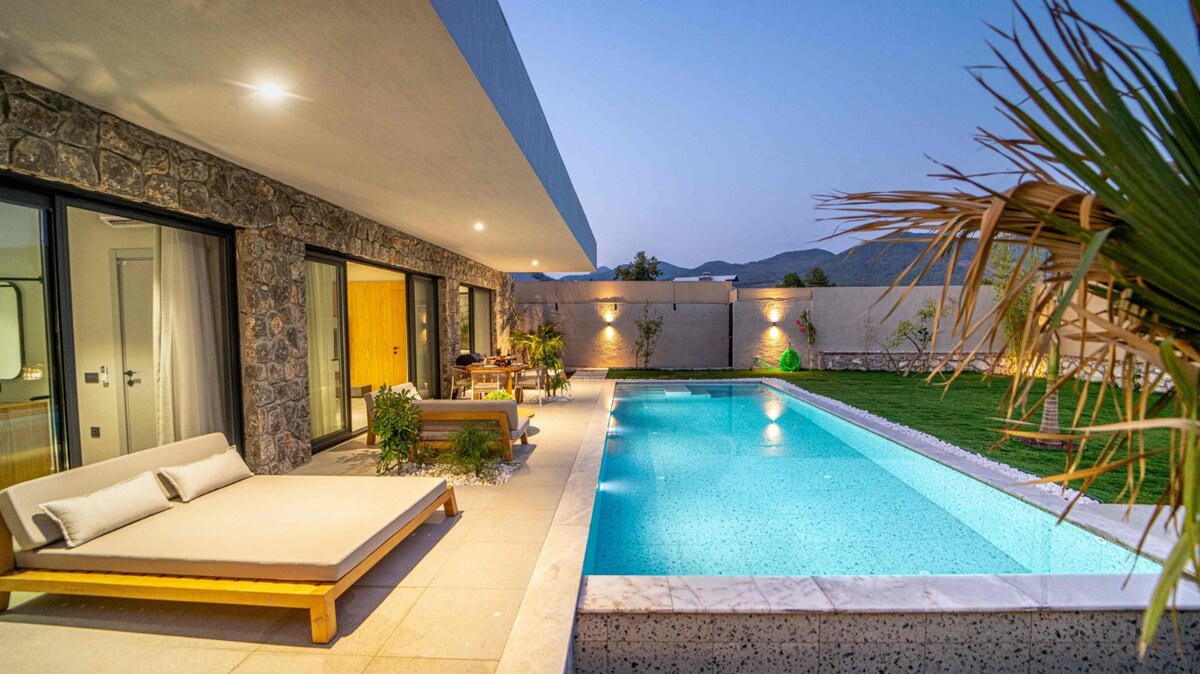 An outdoor area features a lengthy swimming pool surrounded by a smooth stone patio. Lounge chairs are positioned nearby, with tropical plants scattered throughout the space. The villa's modern design is reflected in the large sliding glass doors providing access to the interior.