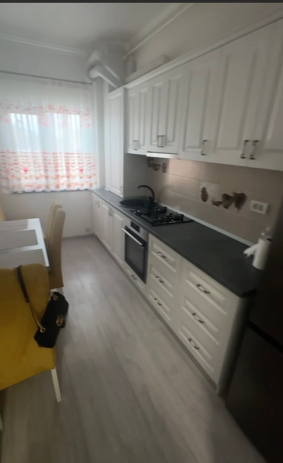 The kitchen features modern white cabinetry with silver handles and a dark countertop. A gas stove is positioned next to a stainless-steel oven. A dining area, with several chairs around a light table, is illuminated by natural light filtering through patterned curtains.