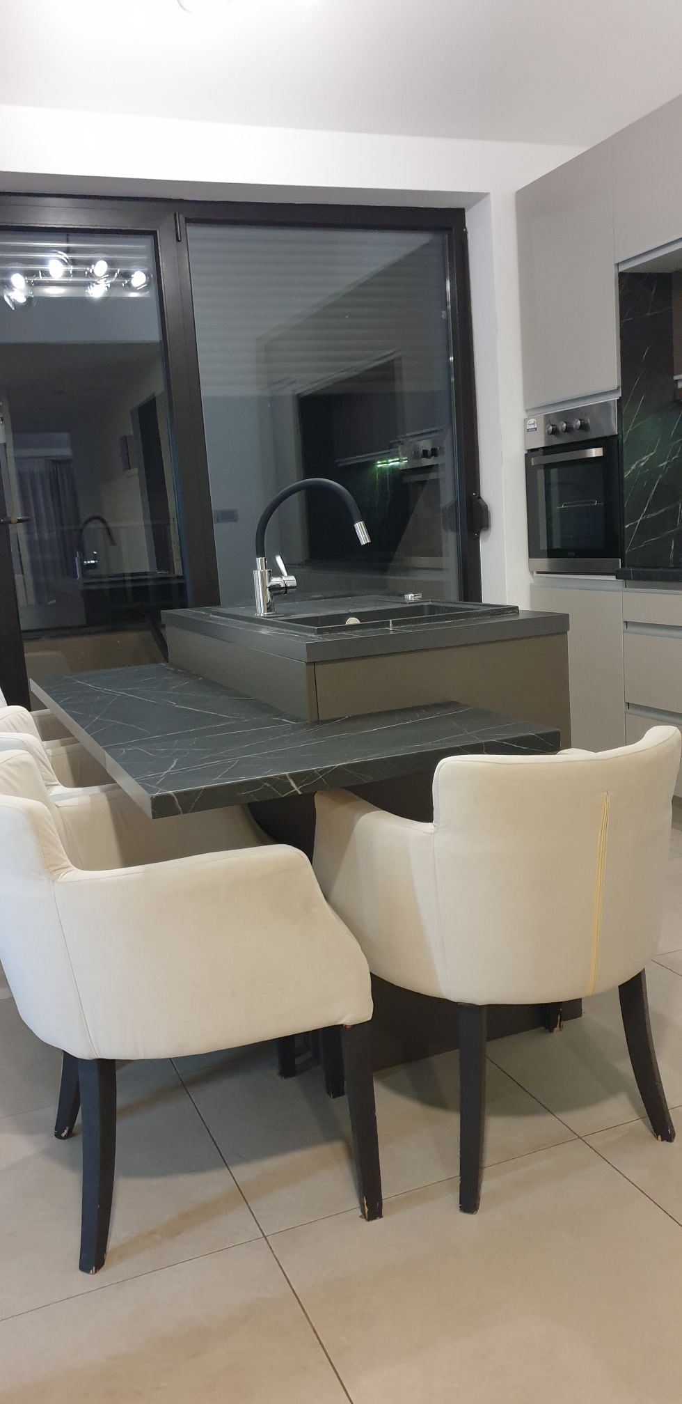 The modern kitchen features a central island with a sleek dark countertop, complemented by four light-colored upholstered chairs. Large windows allow natural light to fill the space, while built-in appliances and minimalist cabinetry add to the contemporary design.
