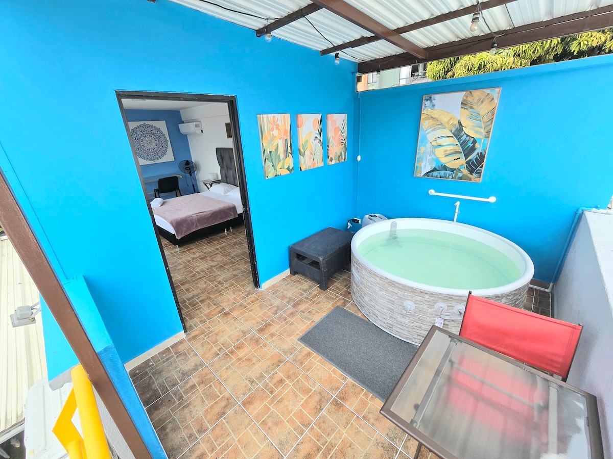 The vibrant blue rooftop area features an inflatable jacuzzi next to a cozy seating arrangement. A glass table and red chairs are positioned beside the jacuzzi. Inside, a spacious bedroom with a queen bed is visible, adorned with colorful artwork on the walls.