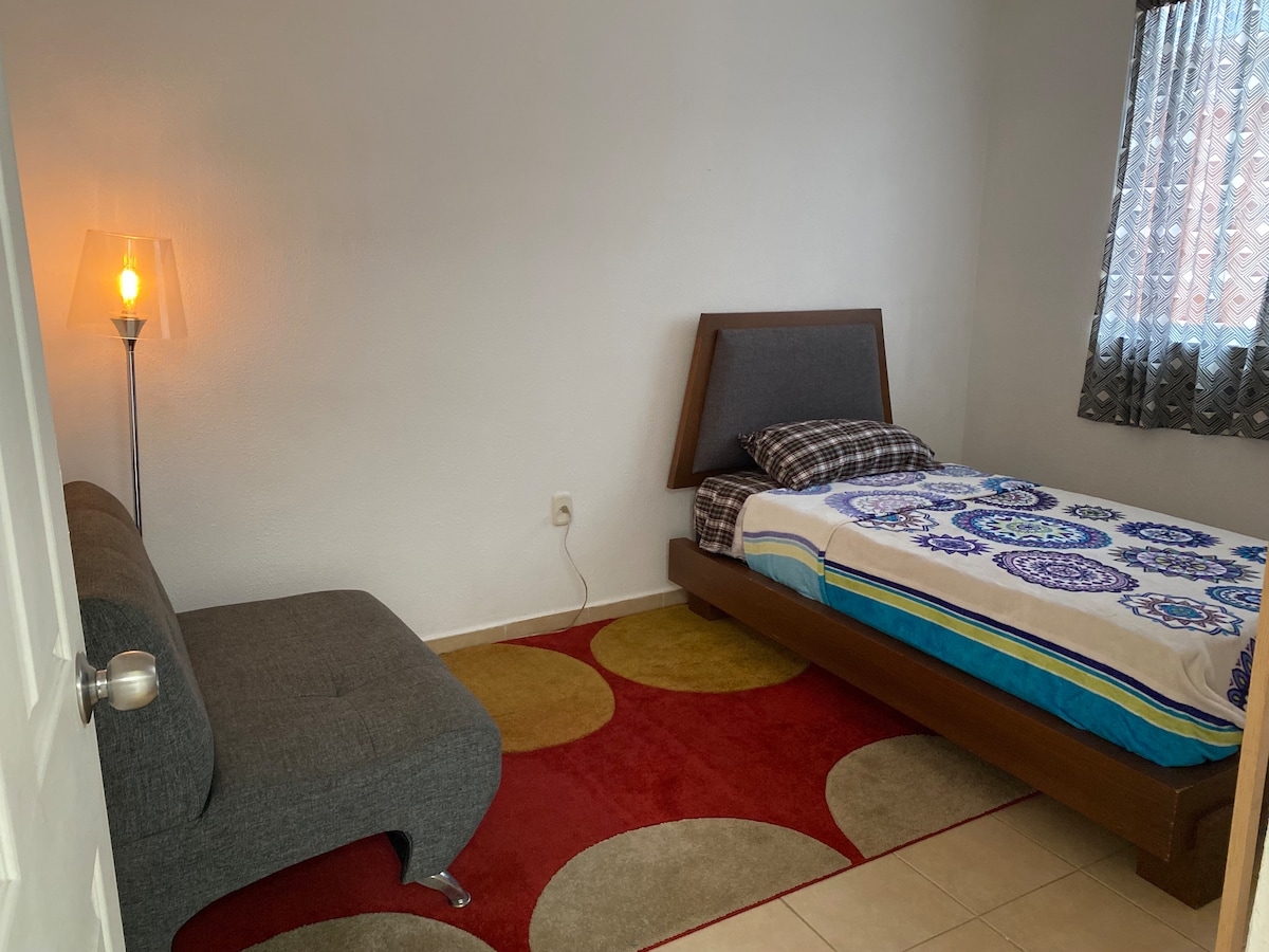 The room features a spacious bed with a patterned blanket, complemented by a cozy seating area with a gray lounge. A lamp with a clear shade provides soft light. Window coverings allow for natural light, adding to the room's serene atmosphere.