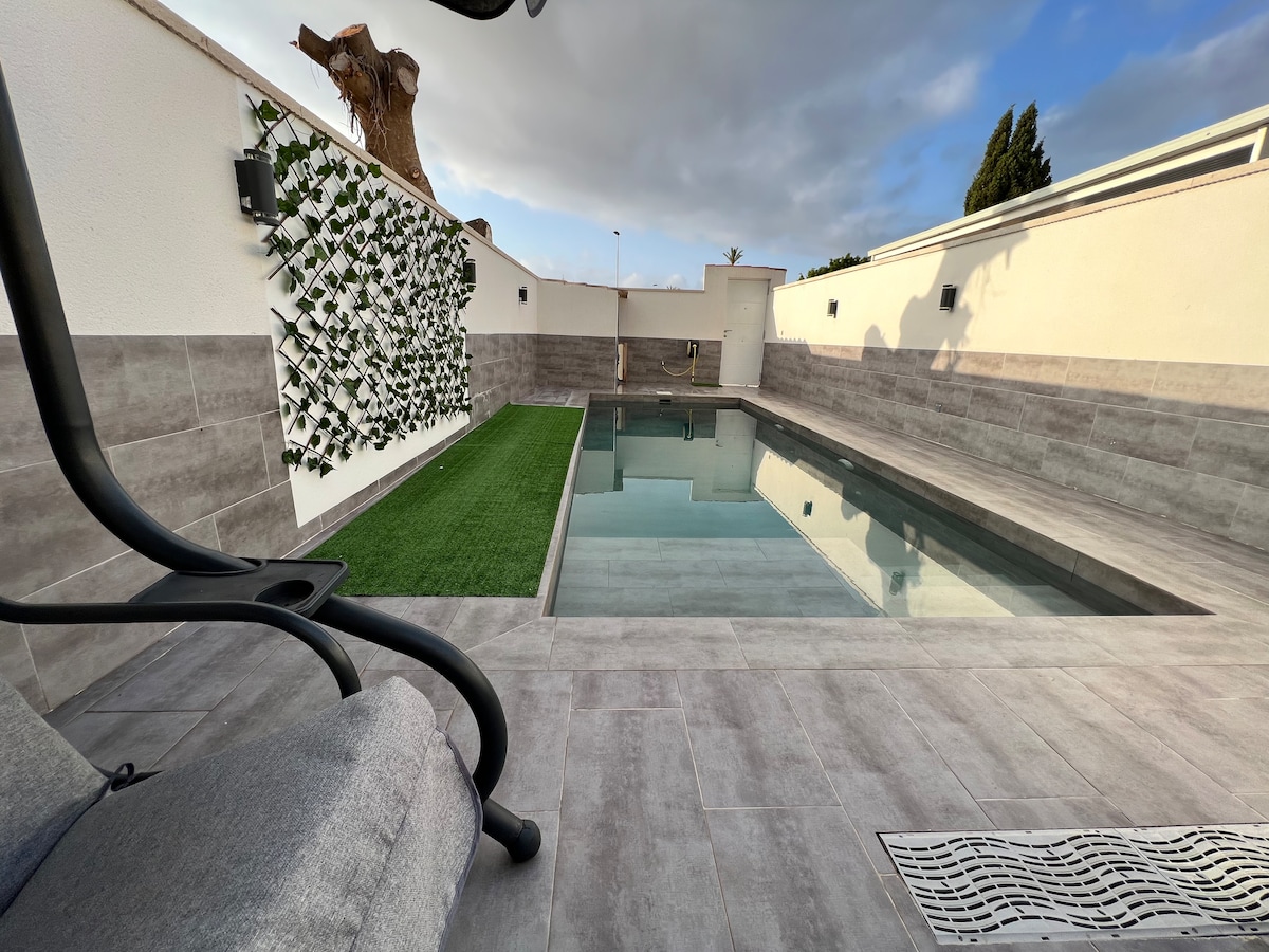 An outdoor area features a rectangular swimming pool lined with smooth tiles. A section of artificial grass is positioned next to the pool, providing a green space. A vertical garden with plants is displayed on the wall, complementing the relaxed atmosphere.