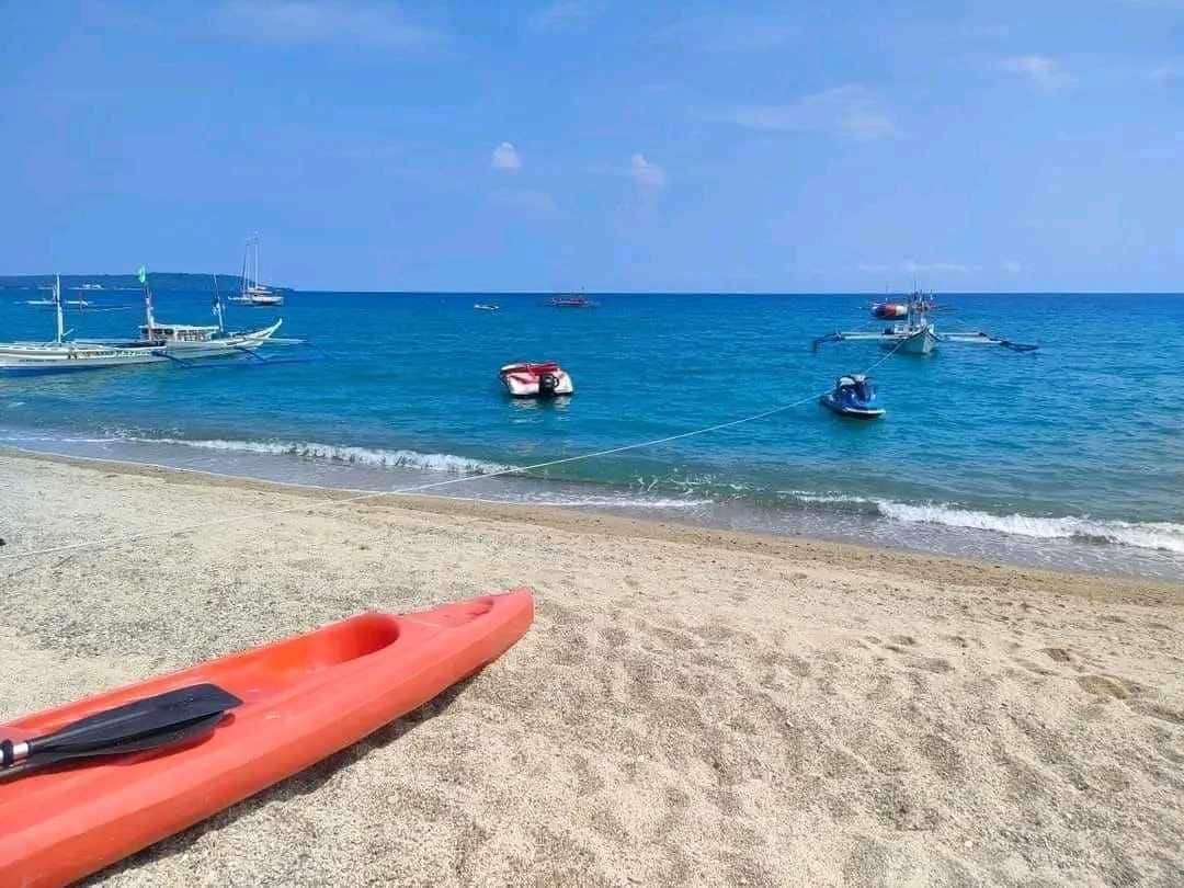 A sandy beach shoreline is depicted, with calm blue waters gently lapping at the edge. Various boats are anchored in the distance, complementing the open sky filled with soft clouds. A bright orange kayak rests in the foreground, adding a pop of color to the serene scene.