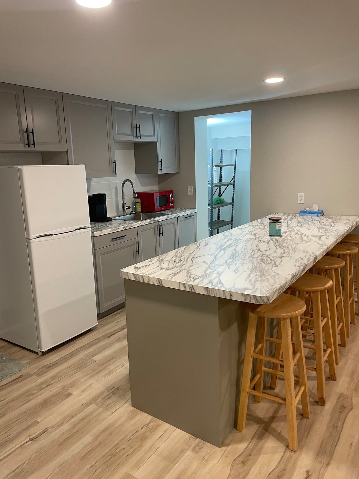 A modern kitchen is displayed, featuring light gray cabinetry and a spacious marble countertop. Barstools line one side of the counter. A white refrigerator, red microwave, and coffee maker are visible. Soft lighting enhances the clean, open layout of the room.