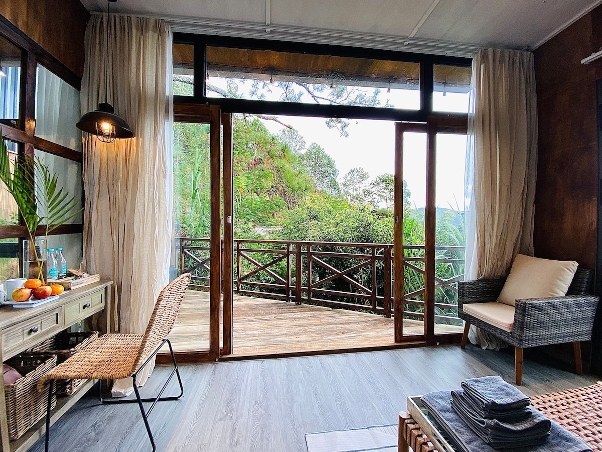 A welcoming living area features large glass doors that open to a wooden deck surrounded by greenery. A comfortable chair and a woven side table are visible, accompanied by a simple desk adorned with fruit and decor. Natural light enhances the space.