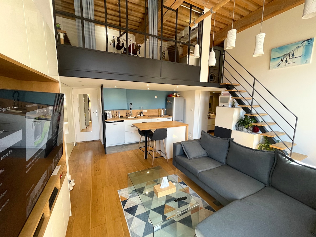 The duplex apartment features an open-plan living area with a modern kitchen and a spacious lounge. A sleek sofa is positioned around a low glass table, with soft natural lighting highlighting the warm wooden flooring. A staircase leads to the mezzanine level, visible above.