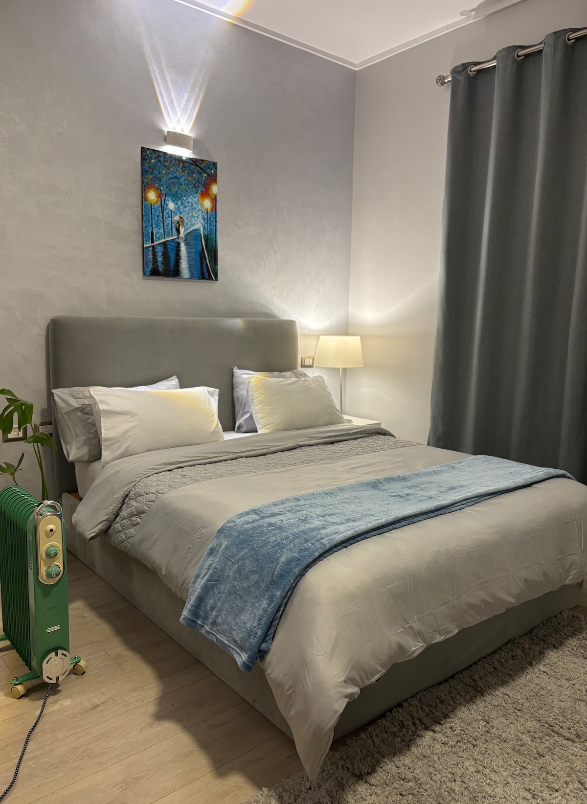 A cozy sleeping area features a comfortable bed with gray bedding, accented by decorative pillows. A soft blanket lies atop the bed. A bedside lamp offers soft illumination, while a piece of art enhances the calming atmosphere. Dark curtains frame the window alongside a small plant.