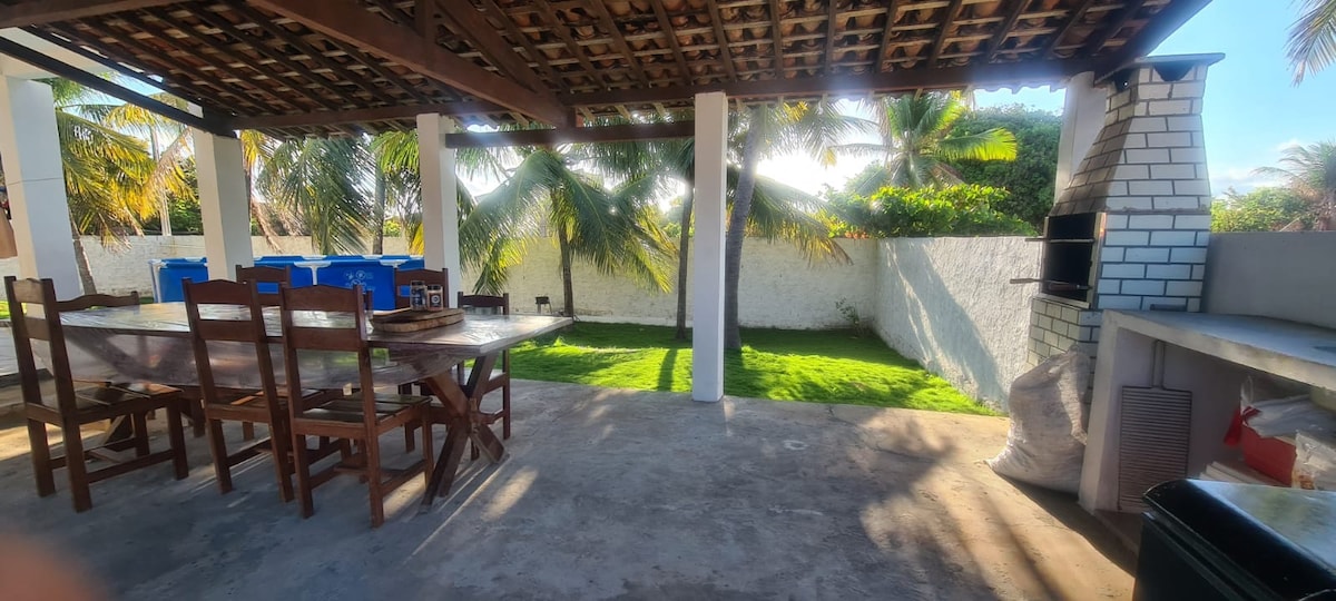 An outdoor dining area features a large wooden table surrounded by several chairs. A barbecue grill is positioned in the corner, with palm trees visible in the background. The space is well-lit, showcasing a grassy area and a white wall in the distance.
