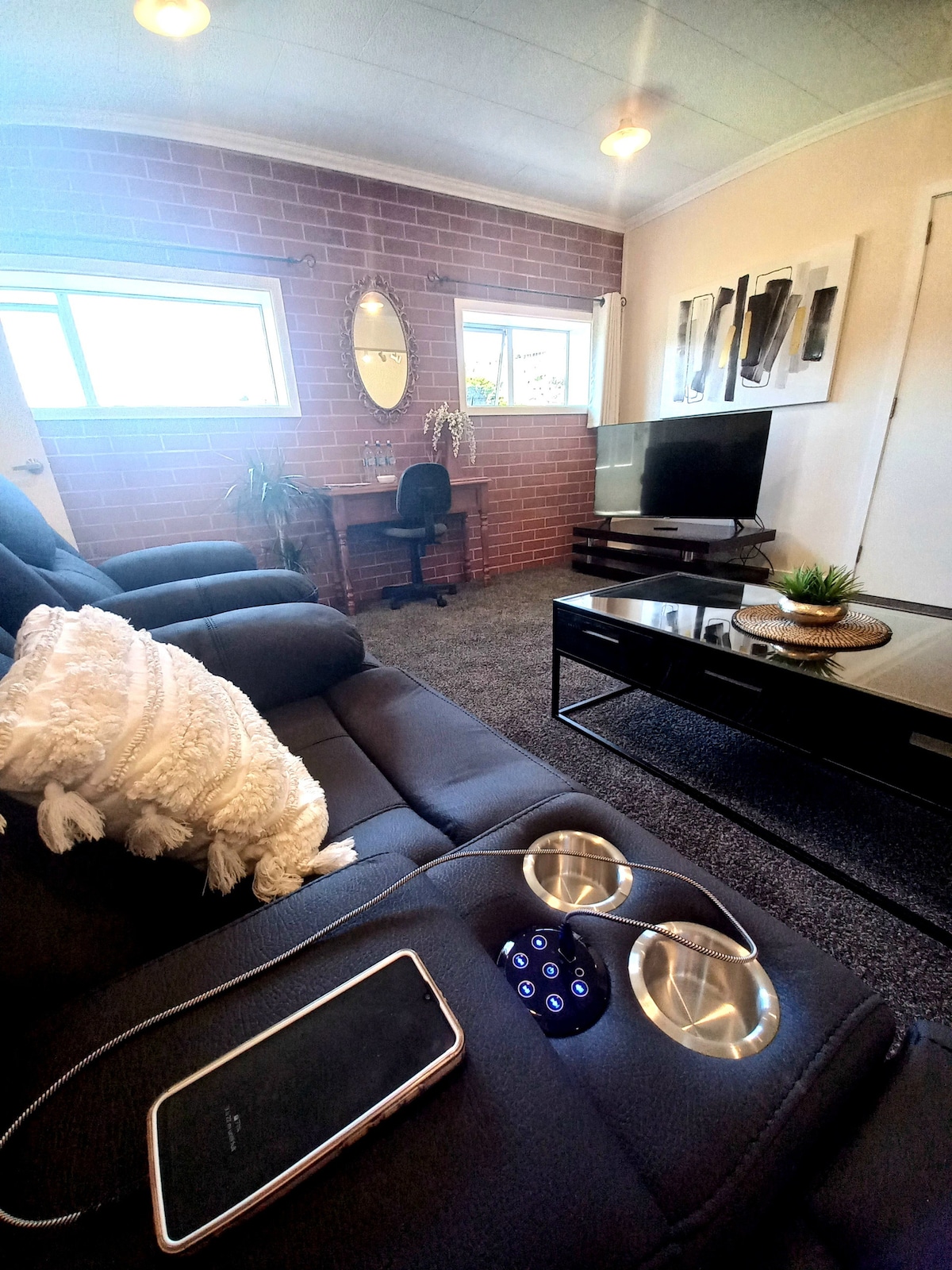 A cozy lounge area features a reclining lounge suite with armrest cup holders. A flat-screen television is positioned on a sleek coffee table. Natural light enters through two windows, illuminating the textured wall and a small desk with a chair in the background.
