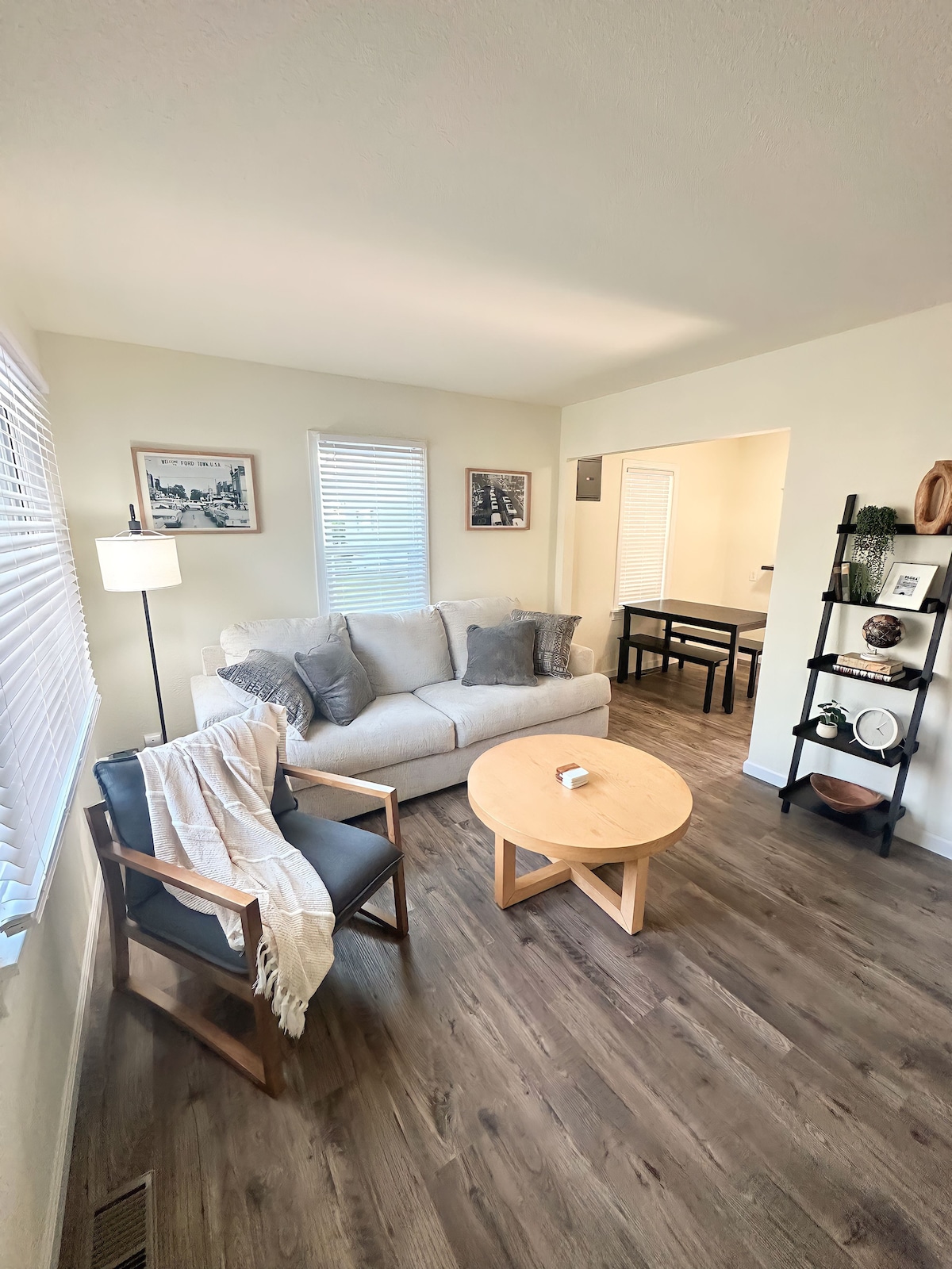 A bright living area features a comfortable sofa adorned with cushions, accompanied by a single chair draped with a blanket. A round coffee table sits in the center, while natural light filters through two windows with blinds, illuminating the wooden flooring.