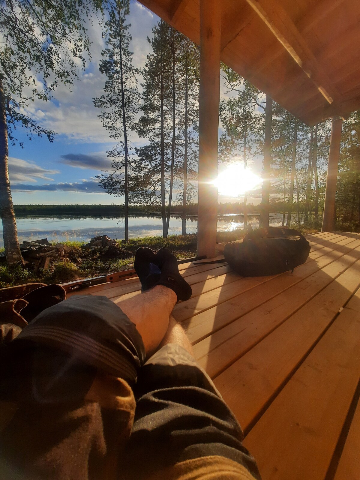 A wooden deck is illuminated by the warm glow of the setting sun, casting long shadows. The calm lake reflects the surrounding trees and sky, creating a serene atmosphere. Personal items are placed on the floor, suggesting comfort and relaxation in nature.