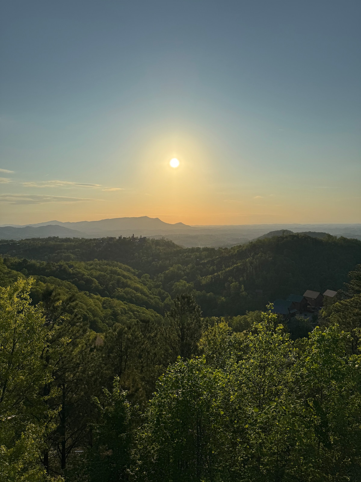 The image captures a serene sunset over rolling green mountains. The sun hangs low in the sky, casting a warm glow over the landscape. Lush foliage in the foreground provides a natural frame, while distant hills create depth, highlighting the tranquility of the mountainous scenery.