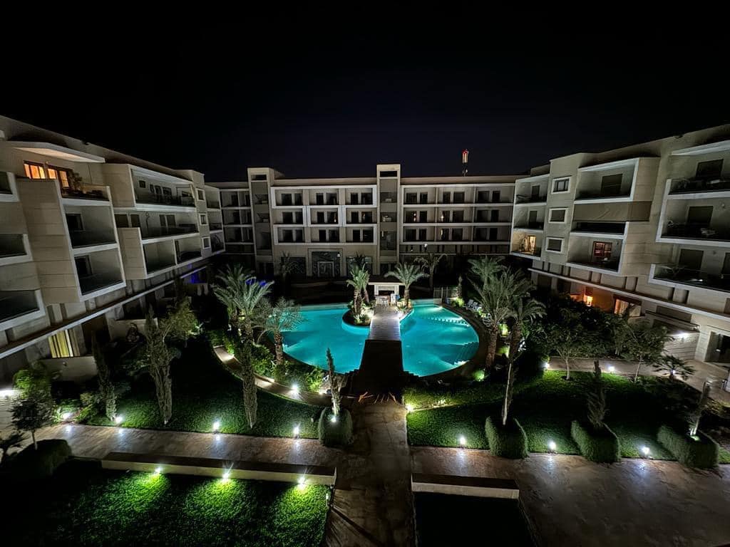 The illuminated courtyard features a central pool surrounded by palm trees and green landscaping. Soft lights highlight the pathways and foliage, enhancing the serene atmosphere of the residential complex at night. Balconies from the surrounding buildings can be seen overlooking the pool area.