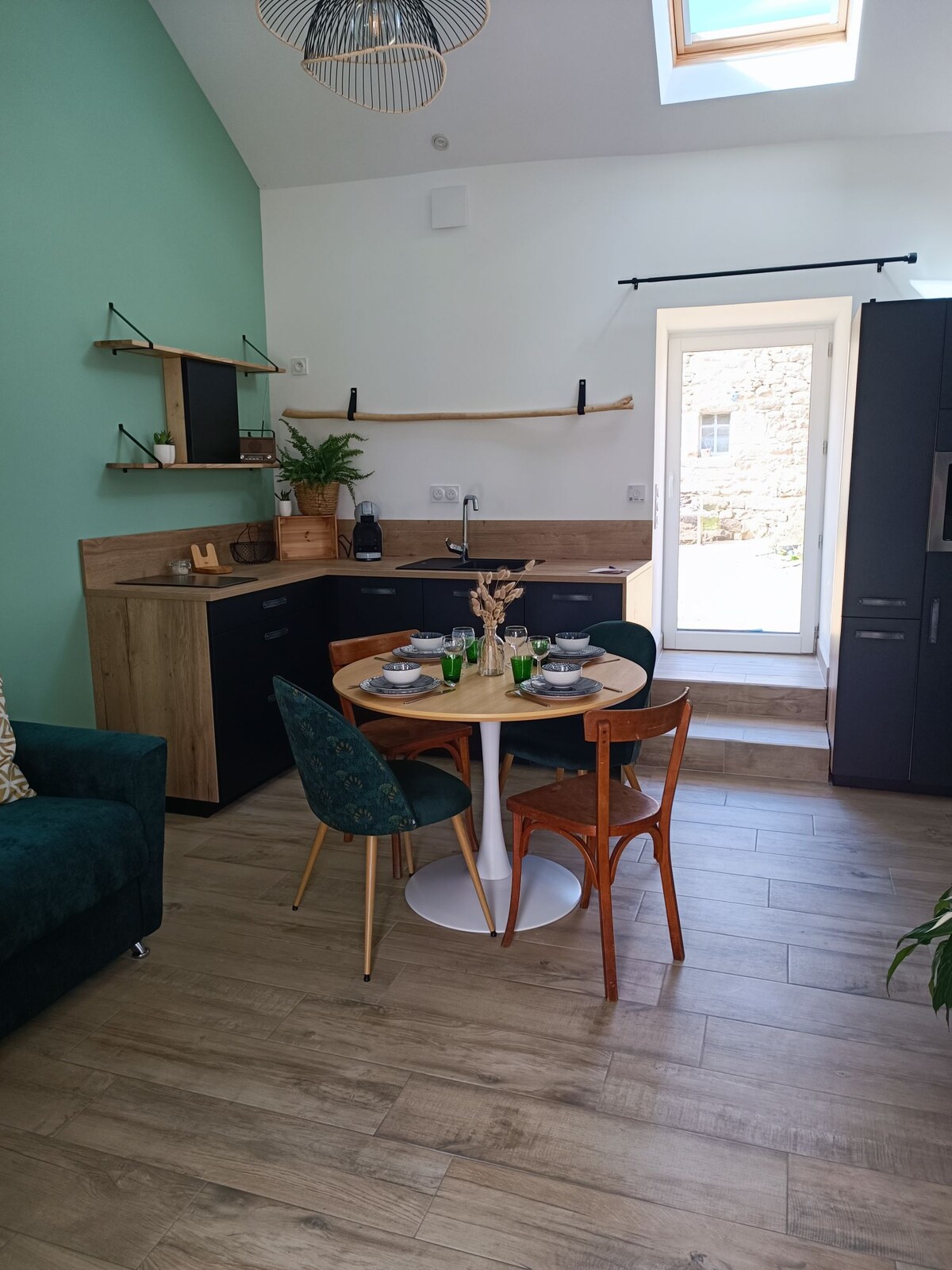 A dining area features a round table set for two, complemented by wooden and upholstered chairs. Kitchen cabinets in muted tones are arranged around the space. A door leading outside is framed by a stone wall, while sunlight filters in from a skylight above.