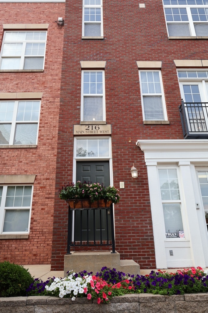 The exterior of a brick townhouse is displayed, featuring a prominent front door with a decorative planter box filled with colorful flowers. Large windows are visible on either side, presenting a welcoming appearance to the home.
