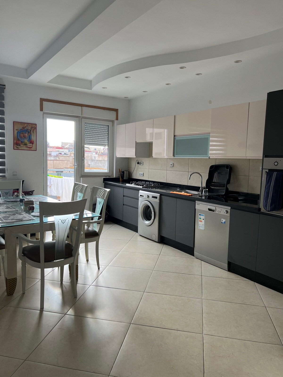 A modern kitchen space features sleek cabinetry in light and dark tones. A dining table with four chairs is situated next to a window, allowing natural light to fill the area. Appliances including a washing machine and dishwasher are visible, adding to the functionality.