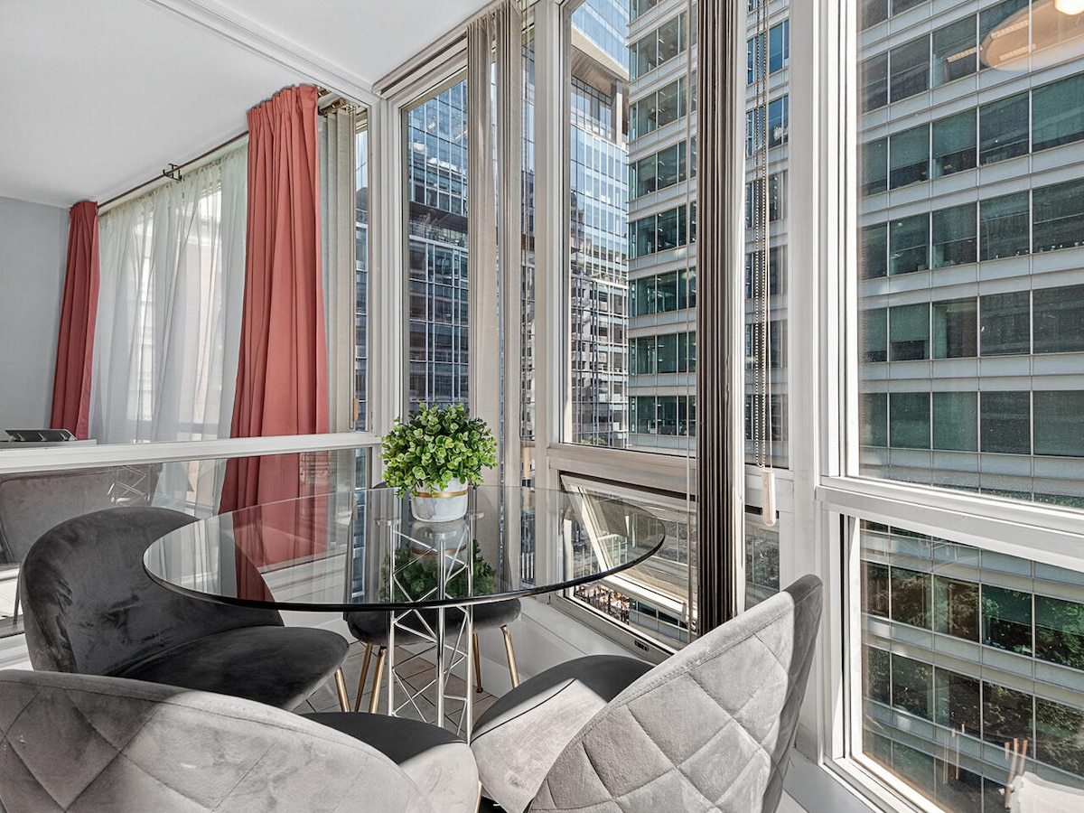 A circular glass dining table is surrounded by plush, grey chairs. Large windows allow natural light to fill the space, revealing views of neighboring high-rise buildings. Sheer curtains softly filter the sunlight, enhancing the modern and inviting ambiance of the dining area.