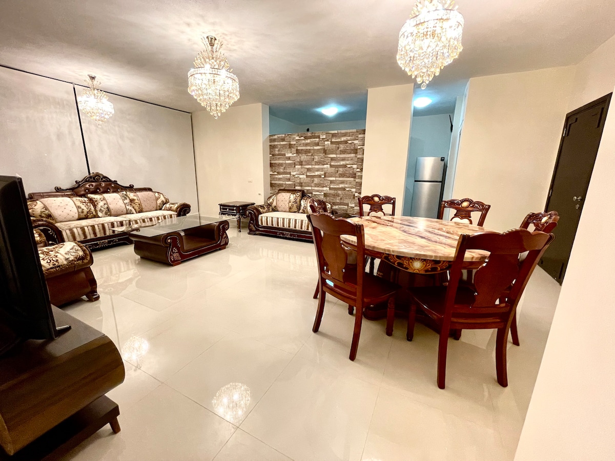 The living room features elegant furniture with intricate woodwork. A large dining table sits in the center, surrounded by comfortable chairs. Crystal chandeliers hang from the ceiling, illuminating the space, while a stone wall adds texture to the backdrop. A modern refrigerator is visible in the corner.