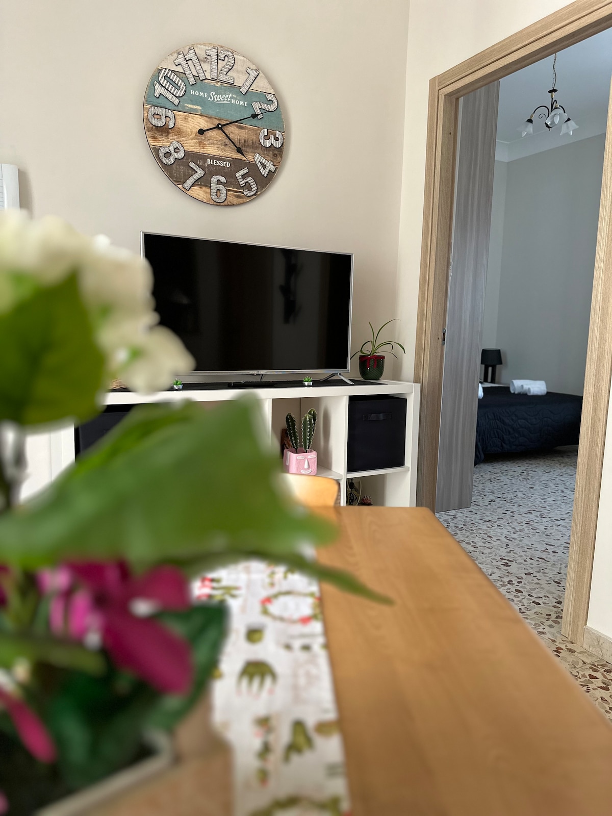 A living area is featured, showcasing a television on a modern white cabinet. A wooden dining table is adorned with a decorative tablecloth and a vibrant potted plant. In the background, a dark-colored bedroom is visible through an open doorway.