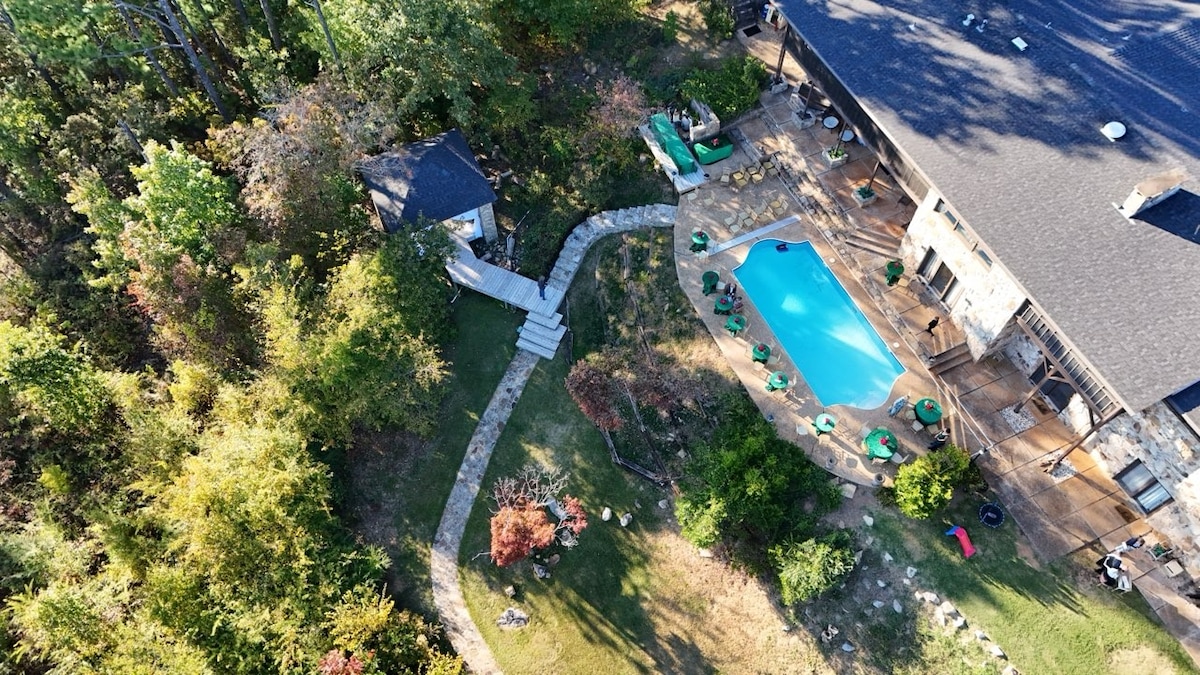 An aerial view showcases a spacious backyard with an inviting pool surrounded by lounge chairs. A winding pathway leads to lush greenery, enhancing the outdoor area. The structure of the property is visible, with a stone exterior and outdoor seating options, ideal for relaxation.