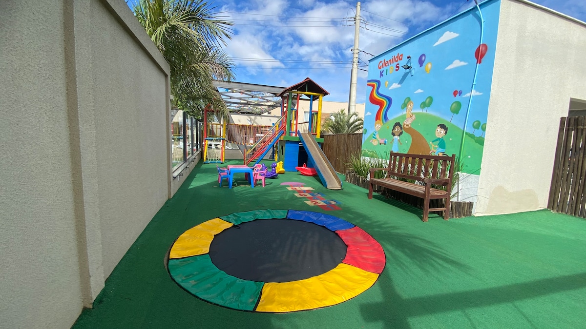 An outdoor play area is visible, featuring a colorful playground structure with slides and climbing features. A bright mural decorates the adjacent wall, showcasing playful designs. Small tables and chairs are scattered on the green turf, providing a friendly environment for children.