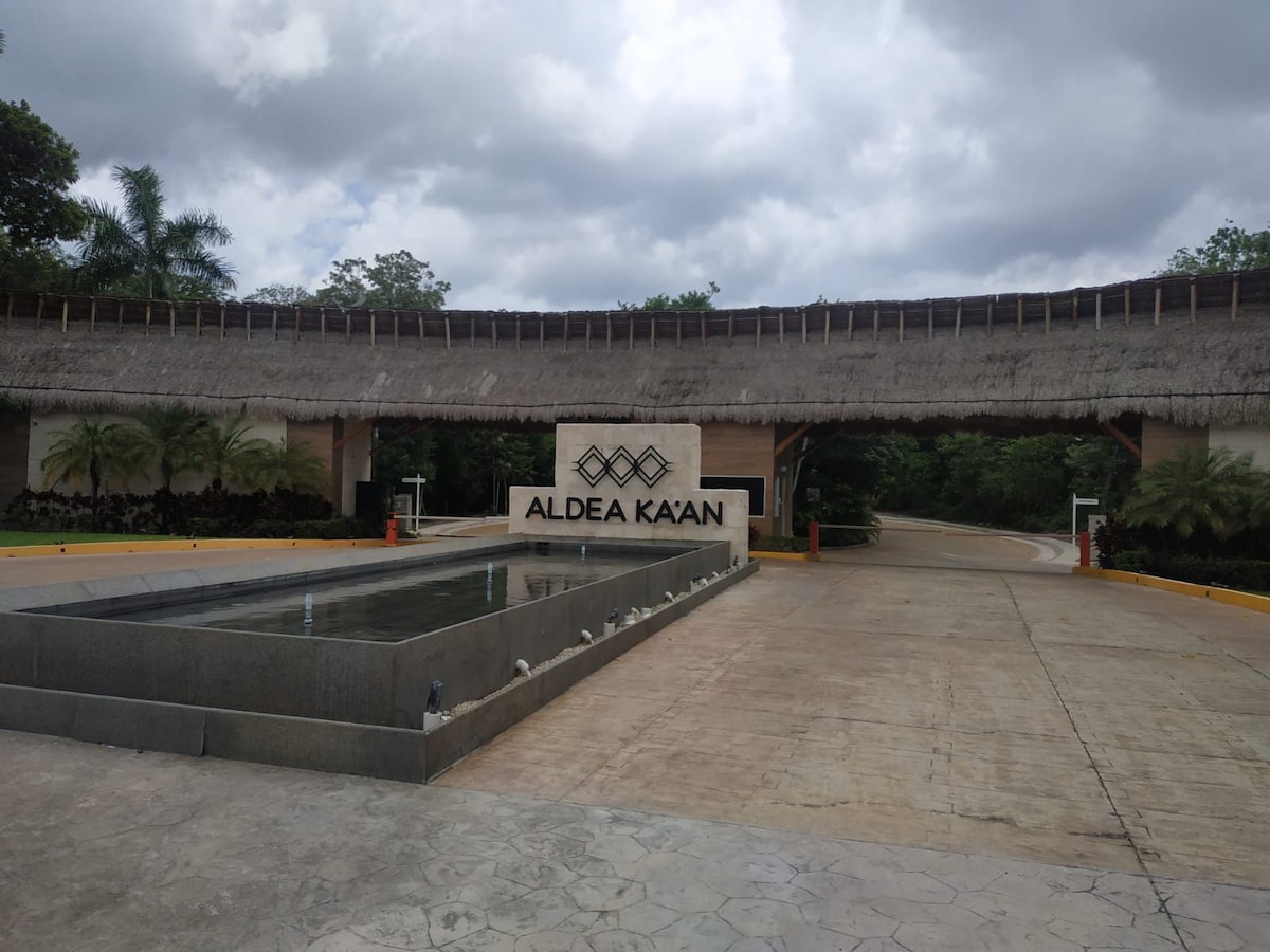 The entrance to Aldea Ka'an is marked by a thatched-roof structure and a water feature. The design is complemented by surrounding tropical plants, and a stone path leads into the community, which is nestled within a natural landscape.