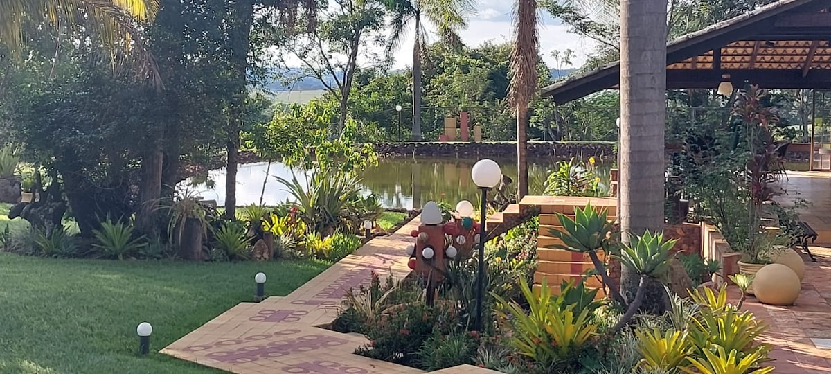 A landscaped garden features a variety of green plants and flowers alongside a serene lake. Decorative lighting elements are arranged among the foliage, and a shaded seating area is visible in the background, creating a peaceful outdoor ambiance.
