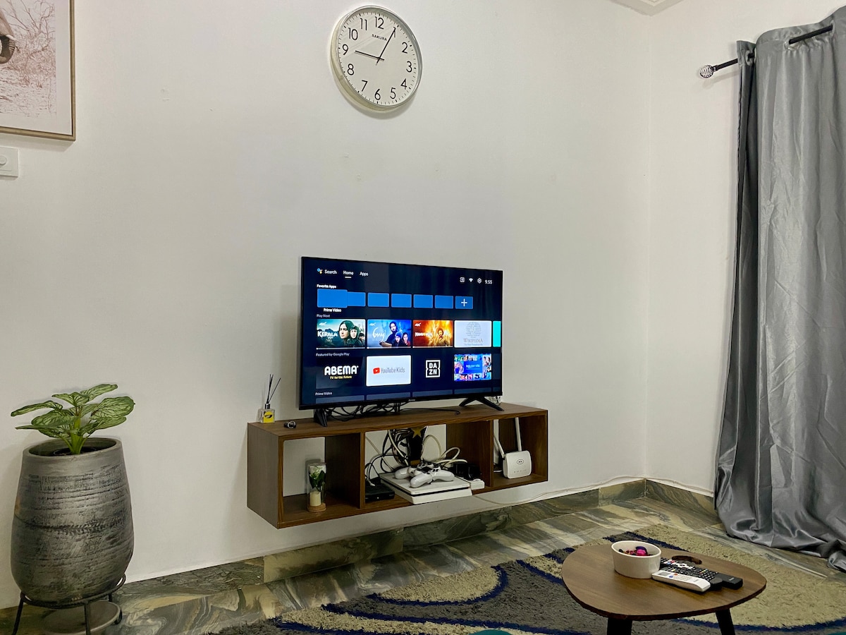 A modern entertainment area features a wall-mounted television with a variety of viewing options displayed. A wooden unit below houses media devices, while a small table sits nearby, adorned with a few decorative items. A potted plant adds a touch of greenery, complemented by wall decor and curtains.