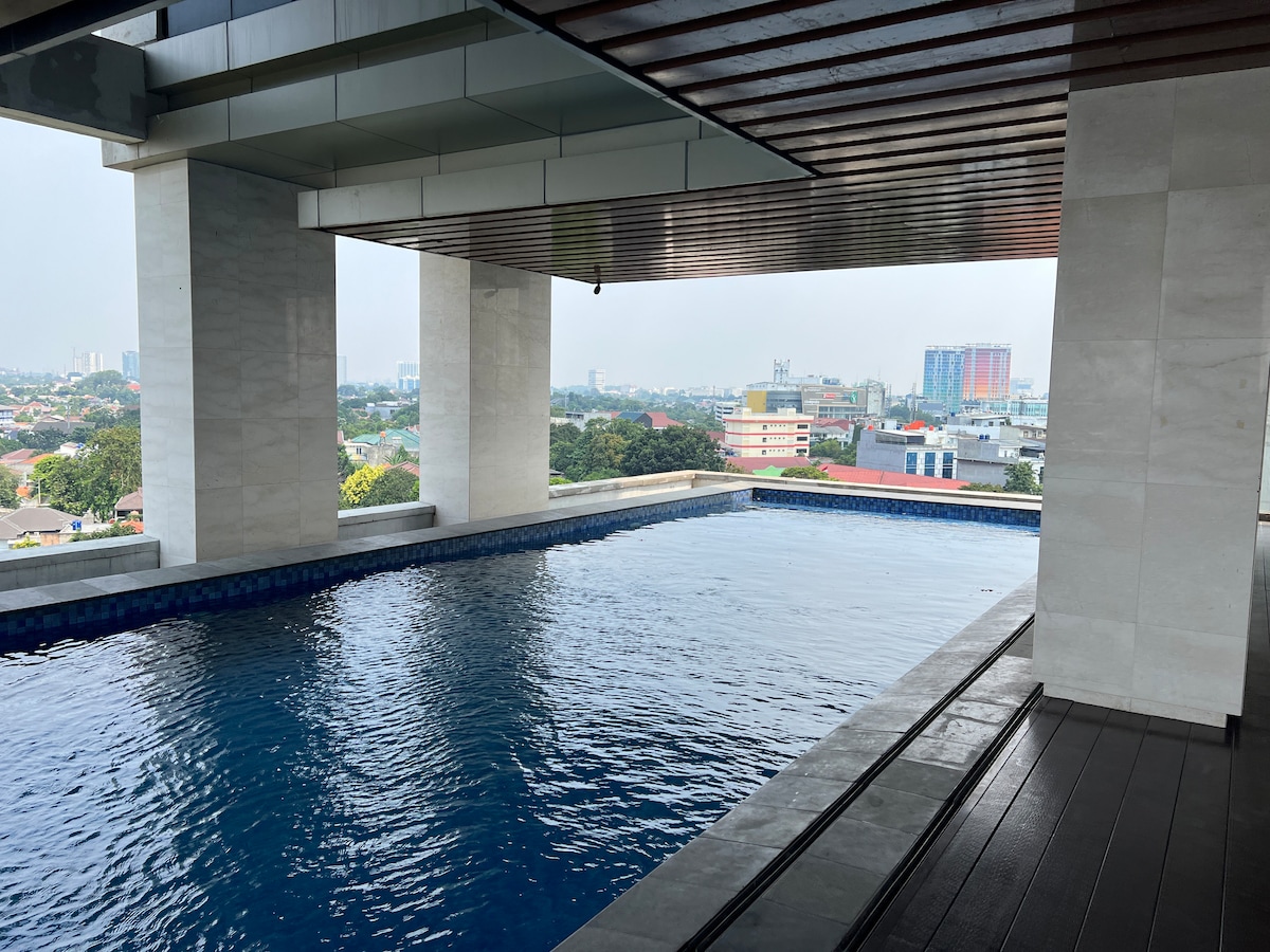 An infinity pool is visible, offering a seamless view of the skyline. Surrounding areas include urban buildings and greenery in the backdrop. The pool's calm water reflects the sky, while the deck is lined with smooth tiles, providing an inviting space for relaxation.
