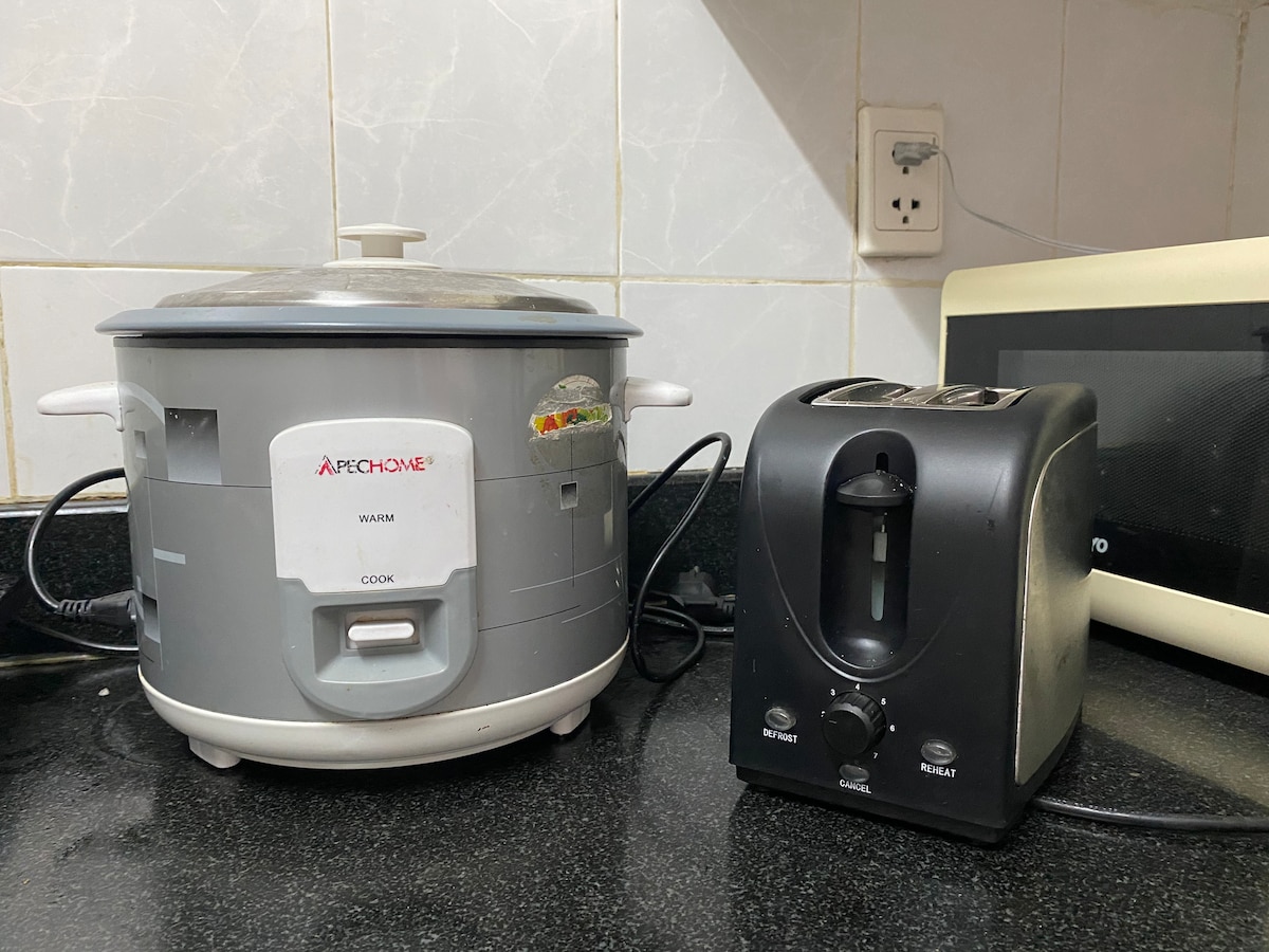 A countertop features a rice cooker and a toaster, both positioned side by side. The rice cooker has a gray exterior with simple controls, while the toaster is black with a single slot. The surrounding area includes white tiled walls and a microwave in the background.
