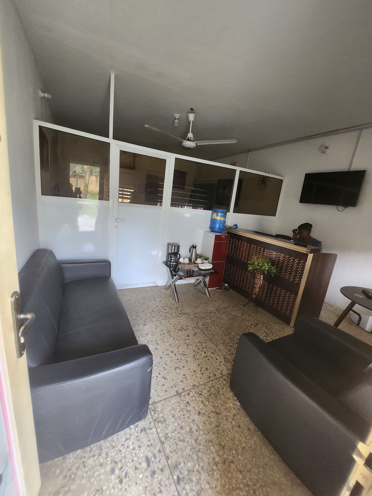 A small reception area features a black sofa and a matching armchair positioned around a coffee table. A fan is mounted on the ceiling, while a water dispenser and a decorative plant add functionality and a touch of greenery to the space. Transparent panels provide separation from the reception desk.
