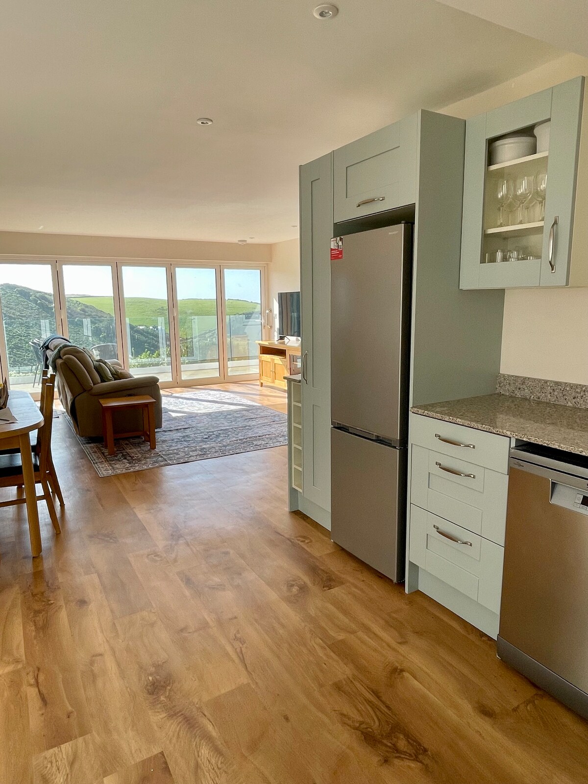 A modern kitchen space features stainless steel appliances, including a refrigerator and dishwasher. Light wood flooring extends through the open area, where a dining table and seating area are visible. Large glass doors allow natural light and provide a view of the surrounding landscape.