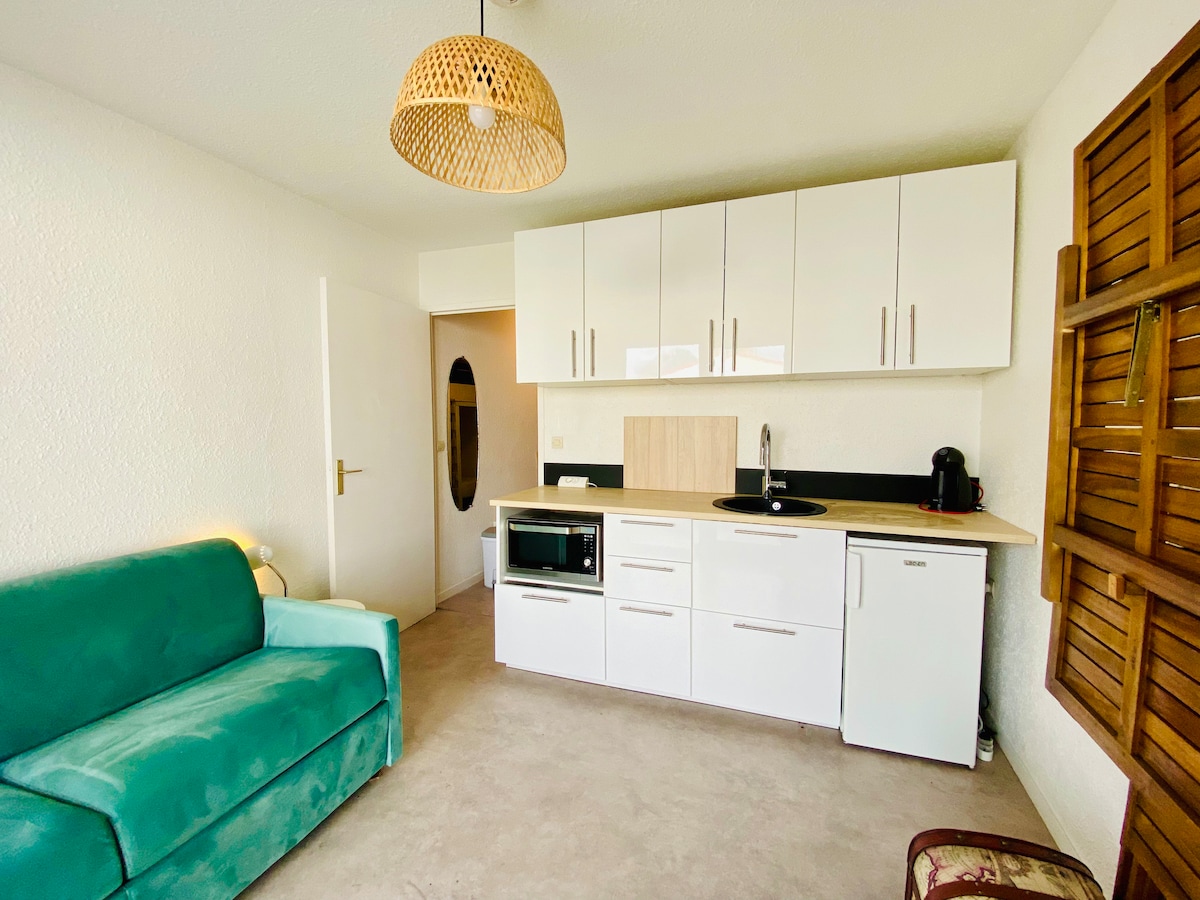 The living area features a bright, open space with a teal sofa and a modern kitchenette. White cabinetry and countertops offer a clean look, while a coffee machine and microwave are visible. Soft natural light is filtered through wooden shutters on the adjacent wall.