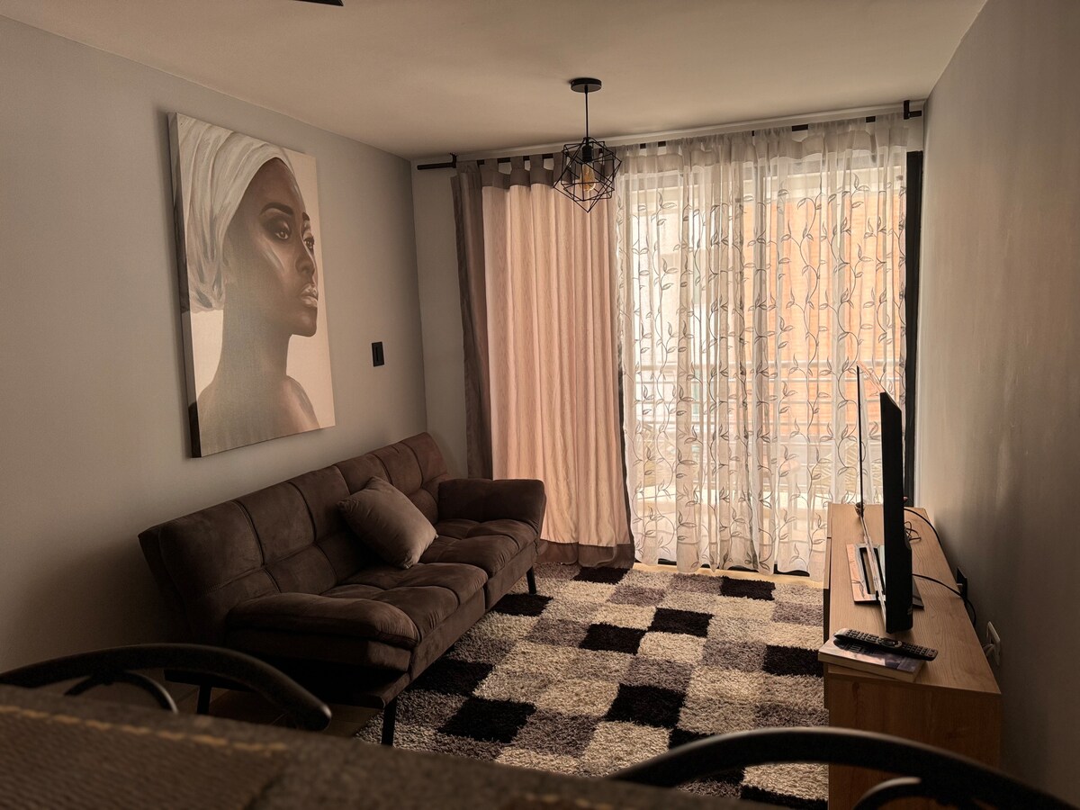 A cozy living area features a plush brown sofa, complemented by a decorative throw pillow. Sheer curtains frame a window, allowing natural light to filter through. A flat-screen TV is positioned on a wooden console, with a patterned area rug anchoring the space.